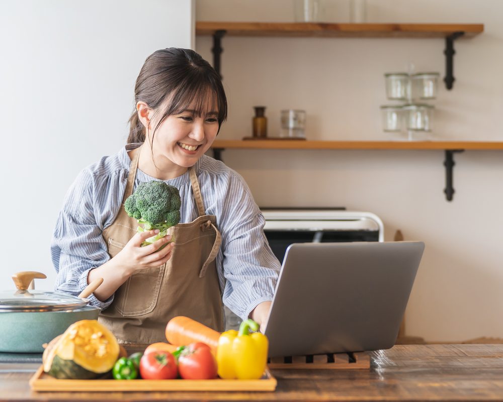 在厨房拿着西兰花、看笔记本电脑查食谱的女士（图库照片，模特摆拍）